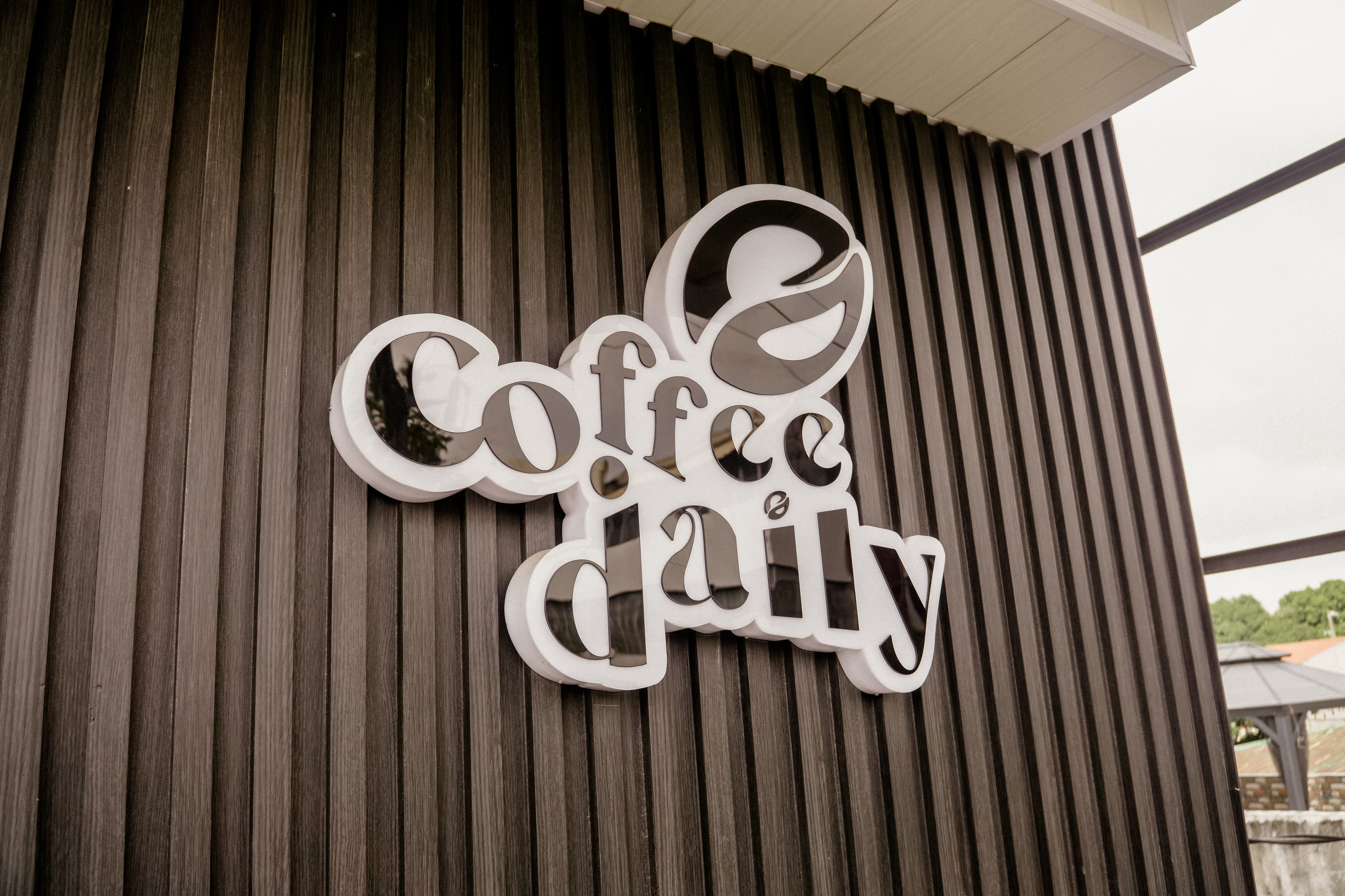 Reflective black Coffee daily sign with bean logo on a dark vertically slatted wall.