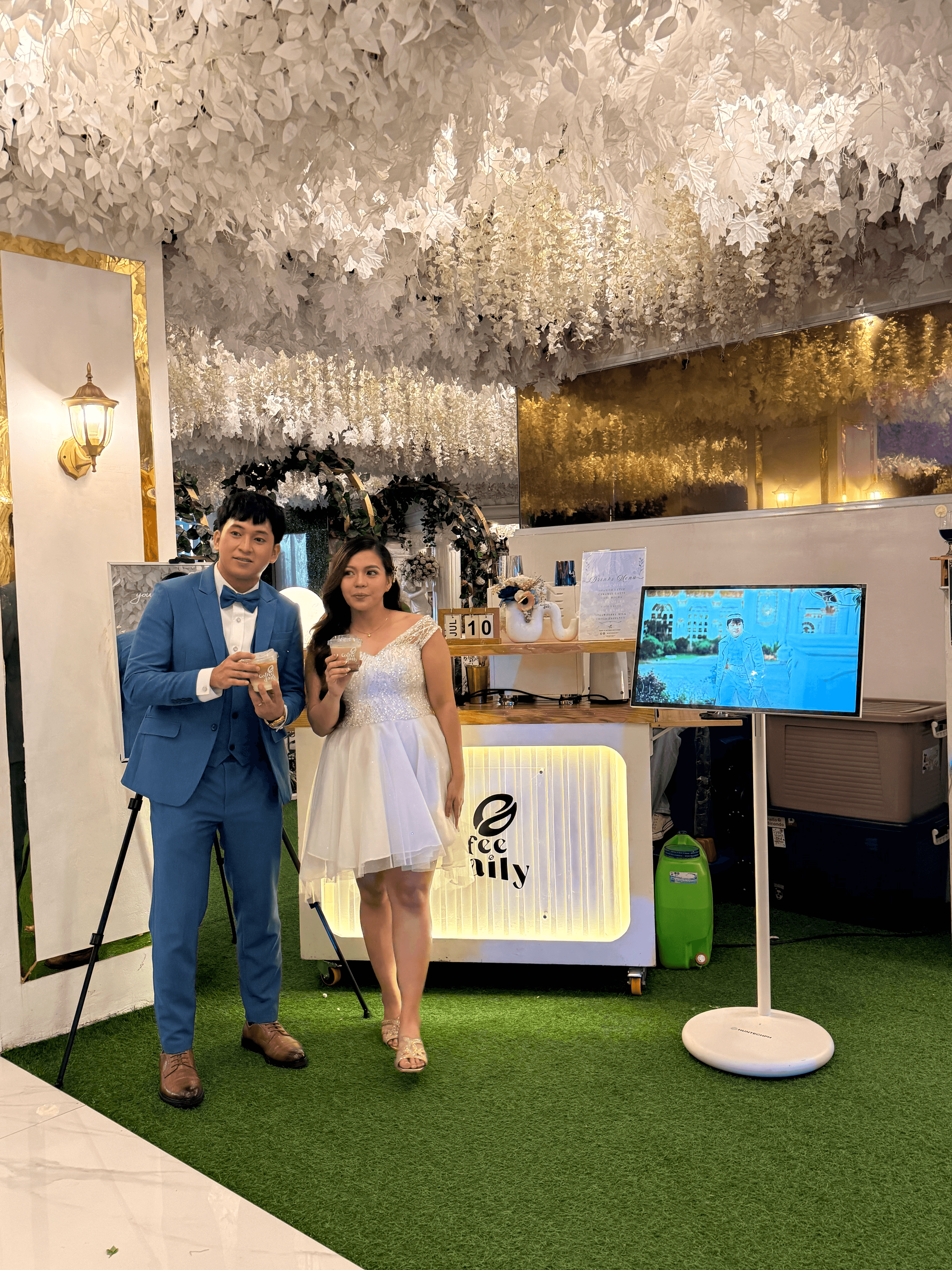 Formal couple posing with drinks at a coffee booth against a white floral backdrop.