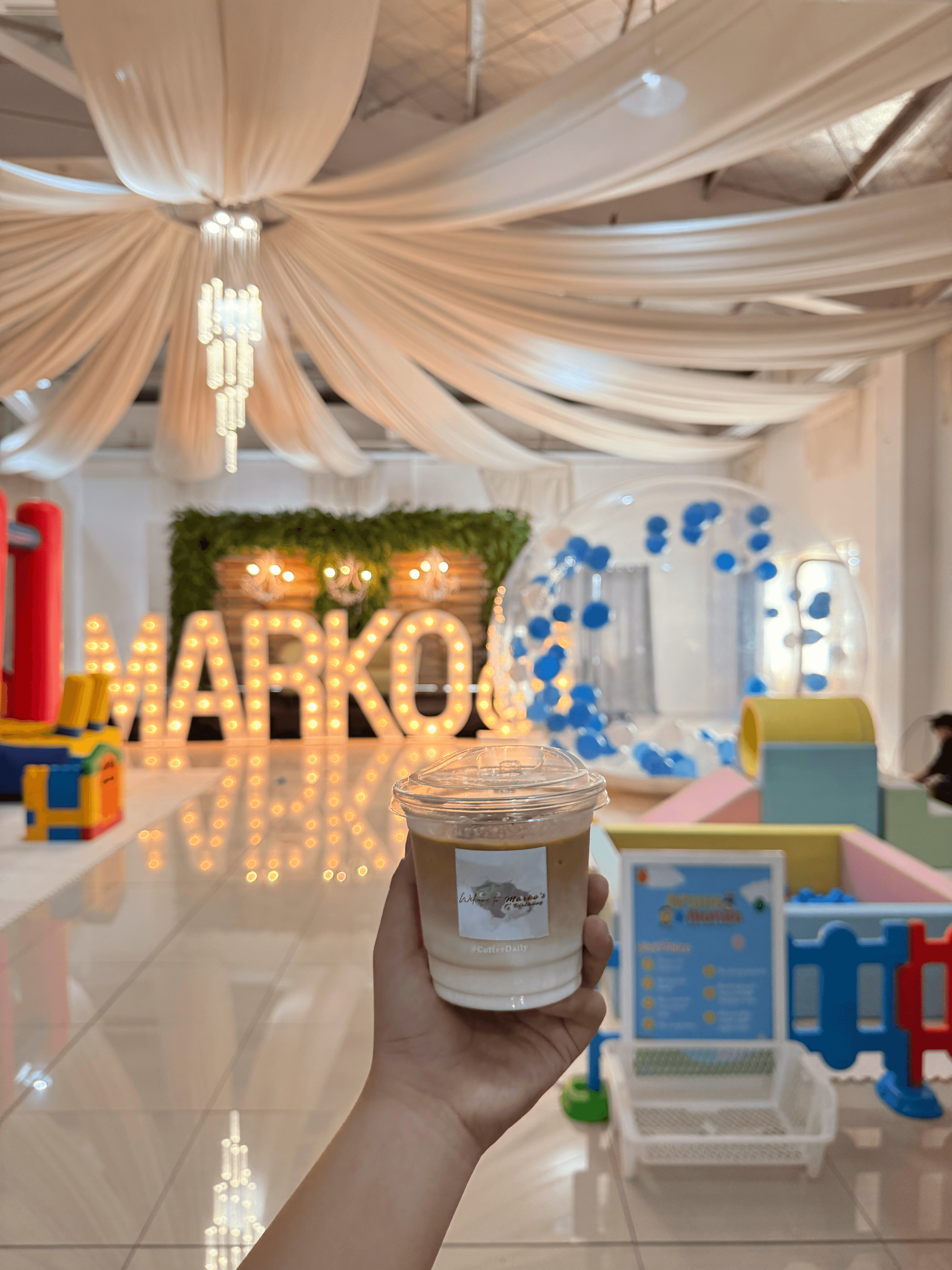 Hand holding iced coffee before a lit 'MARKO' marquee sign and white fabric drapes.
