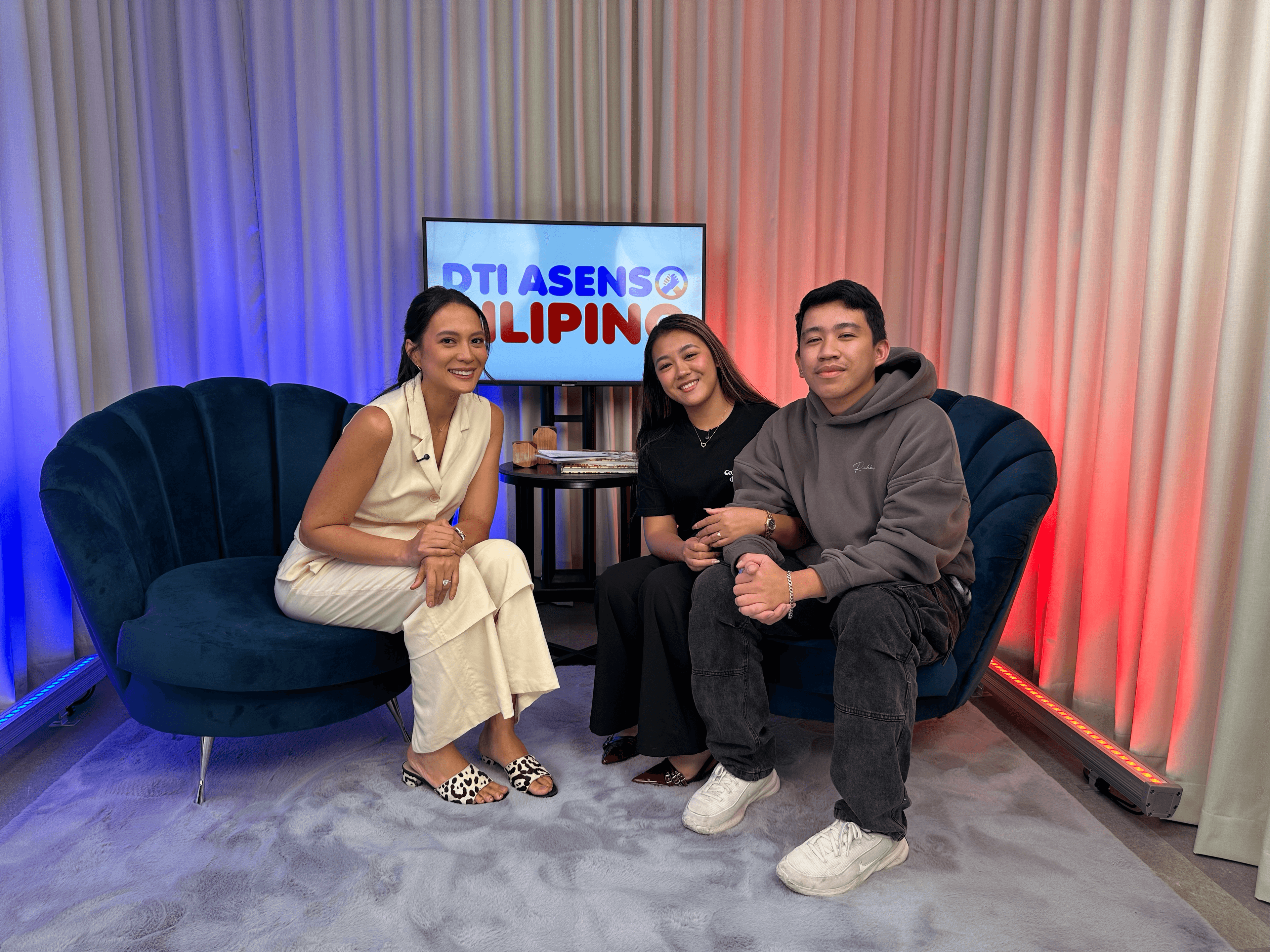 Three people sit on blue chairs in a studio with a DTI ASENSO PILIPINO screen.