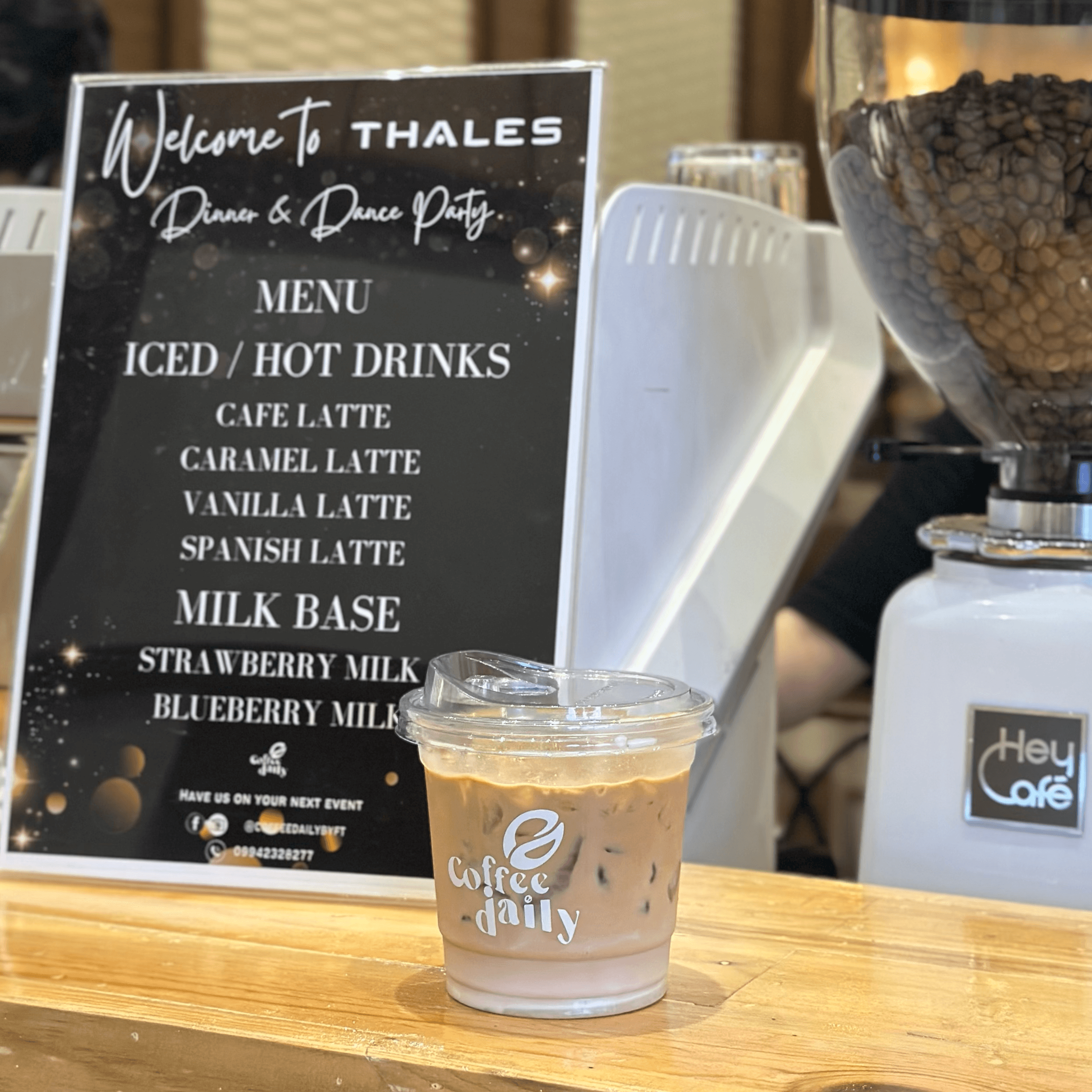 Iced coffee cup on a wooden counter beside a Thales event menu and coffee grinder.